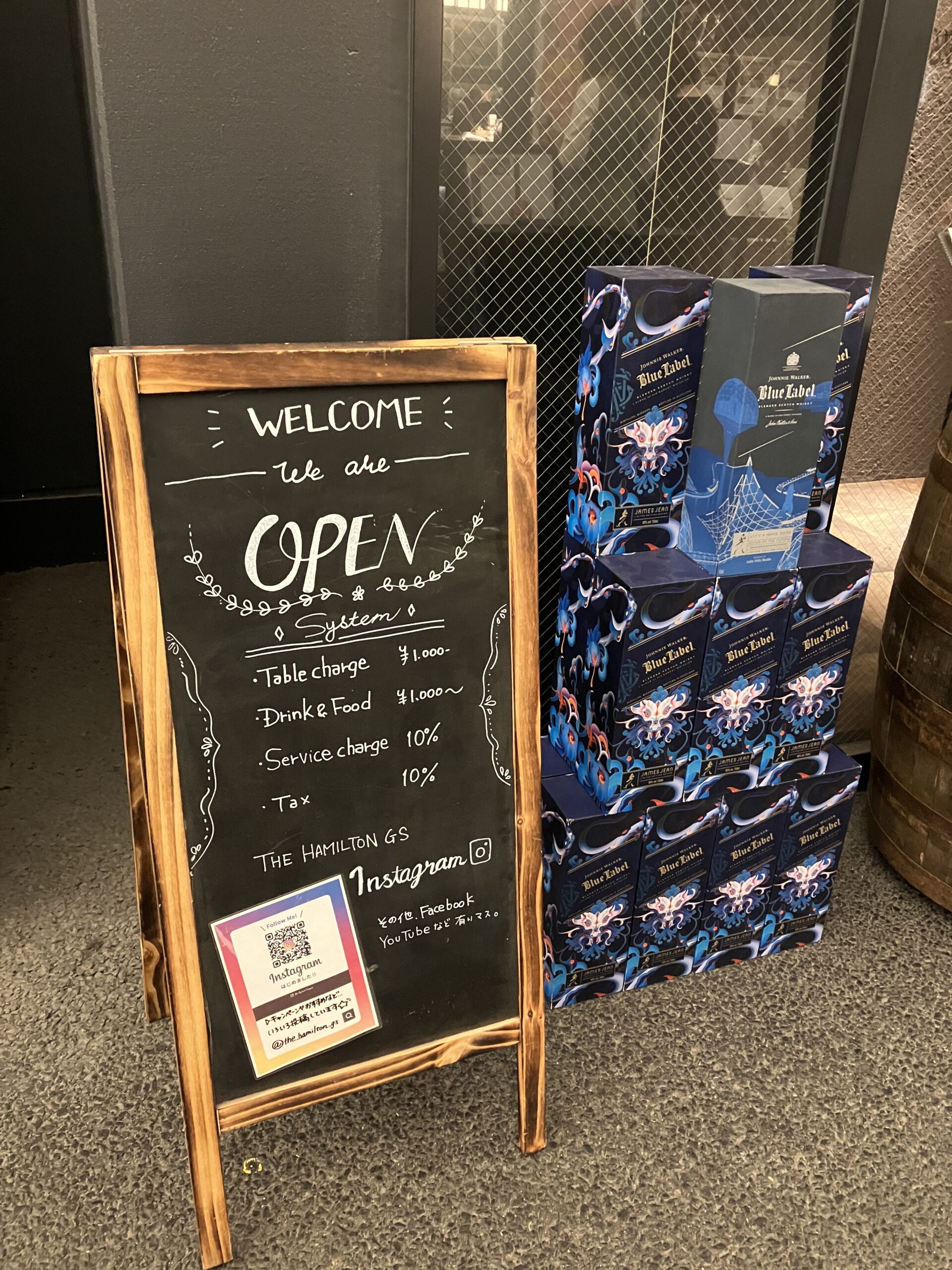 「THE HAMILTON GS」の入り口付近の写真。立て看板が立っている。開店していることとInstagramのQRコードが掲示されている。